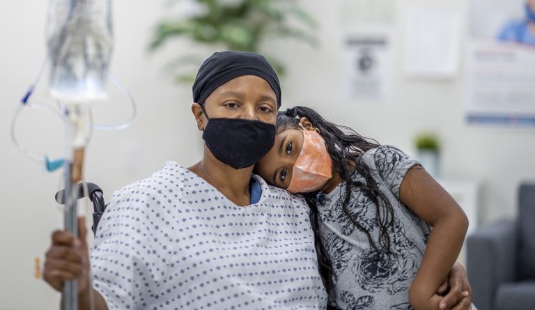 a woman with cancer holds an IV and embraces with her granddaughter; both are wearing masks