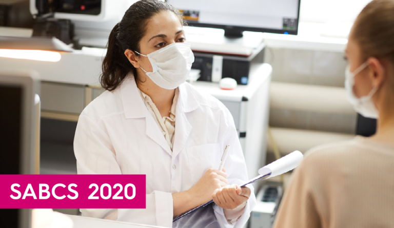Doctor wearing a lab coat and mask holding a clip board speaks to a patient