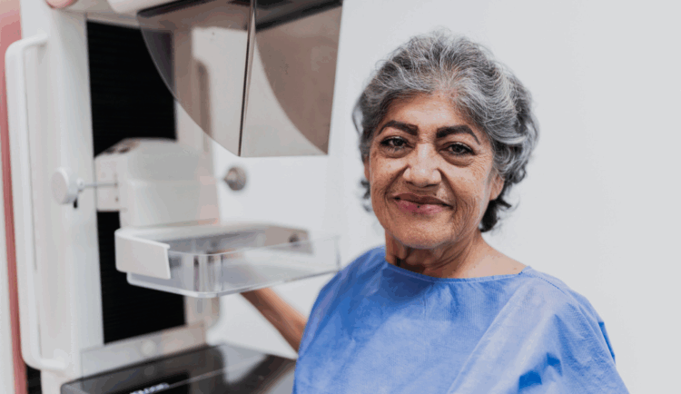 a woman prepares for her mammogram, she stands in front of the machine and is smiling at the camera wearing a blue gown