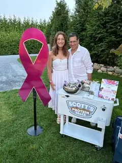 lori levine and her husband scott outside next to a pink ribbon and a stand of scott's protein balls